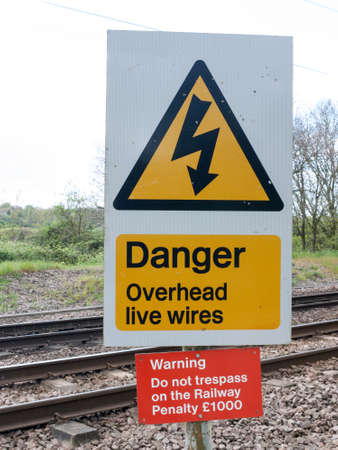 A Railway Safety Sign Saying Danger Overhead Live Wire Warning Do Not Trespass On the Railway Penalty Â£1000 in Black, Yellow, Red, and white with Track in Background in Spring in UKのeditorial素材