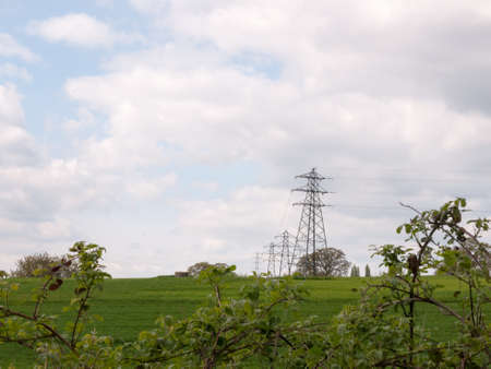 a shot of far away field green and with several lined up electrical wire metal frames standing and with a beautiful background of white and blue skies during the day of spring and trees upfront near train tracksの写真素材