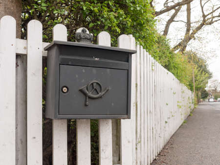 an interesting and unique black post box outside locked with an icon and attached to a white fence on a path and with hedge in spring clear delivery homeの写真素材