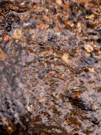 the rippled surface of running water with multicolored natural pebbles below the water surface and some sticking out shining and glistening as water travels over and down stream in the day lightの写真素材