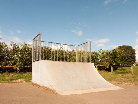 An Empty and Unused Ramp Half Pipe at the Skate Park in the Country Park in the UK Shining in the Sunlight of the Day and with A Shrub Hedge Behind the Bars Protecting it Fenced in the Spring in the UKの写真素材