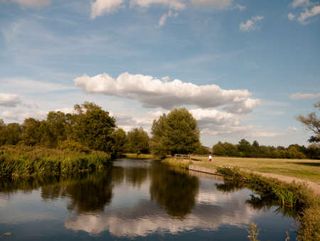 long cloud and blue sky reflected in river with trees in the country lady walking; UKのeditorial素材