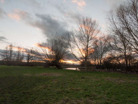 sunset moody sky winter autumn over green field with trees water; essex; england; ukの写真素材