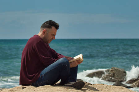 man sitting on the sand reading a book on the seashore with the sea in the backgroundの写真素材