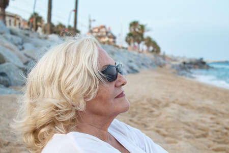mature woman in sunglasses sitting alone on the beach by the sea in a white dressの写真素材