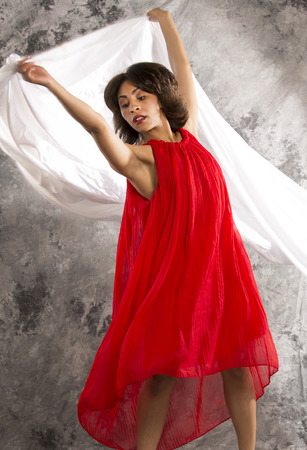Attractive young, African American, female dancer, in red dress, in dramatic turns displaying white fabric against a grey background. Vertical image.の写真素材