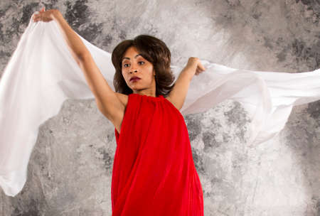Attractive young female dancer, representing diversity in dance, in red dress, displaying white fabric against a grey background.の写真素材