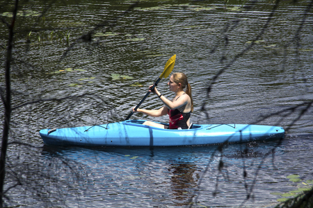 Young woman in blue kayak, gently paddling left on Mud Pond in Sunapee, New Hampshire, framed by tree branches on a sunny day, horizontal image.の写真素材