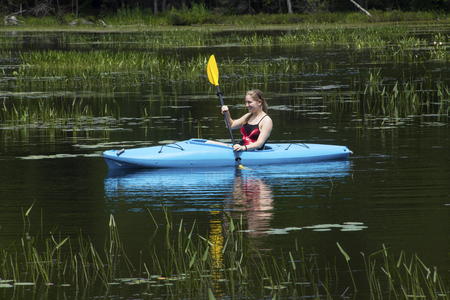 Young woman in blue kayak, paddling left through water plants on Mud Pond in Sunapee, New Hampshire, on a sunny day, horizontal image.の写真素材