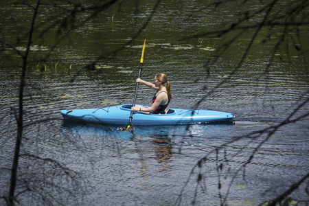 Young woman in blue kayak, paddling left on Mud Pond in Sunapee, New Hampshire, on a sunny day, seen through web of tree branches, horizontal image.の写真素材
