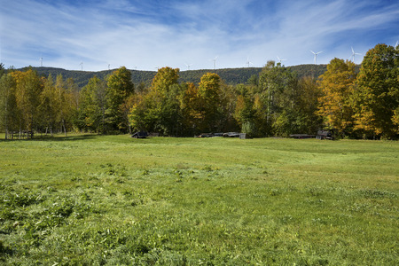 View of a windfarm with a dozen windmills on a high ridge above green grass of a pasture, with fall foliage and a blue sky on a sunny day in northern Maine, near Roxbury.の写真素材