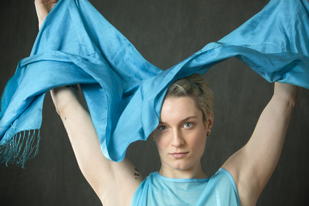 Head and shoulders portrait of strong young woman dancer with short blonde hair, in a white leotard and blue cape, spreading a blue scarf.の写真素材
