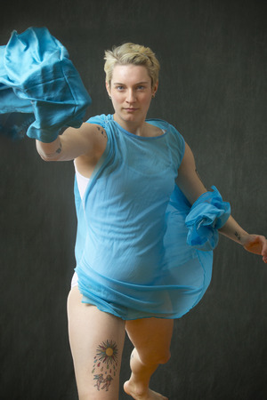 Active young woman dancer with short blonde hair, in a white leotard with a blue cape, stepping forward while throwing a blue scarf in the studio.の写真素材