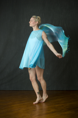 Attractive young woman dancer with short blonde hair, in a white leotard with a blue cape, swinging a blue scarf in the studio.の写真素材