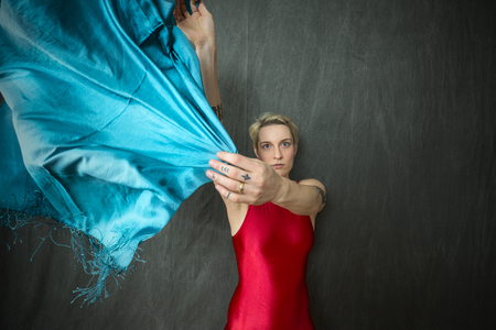 Upper body portrait of active young woman dancer with short blonde hair, in a red unitard waving a blue scarf in the studio.の写真素材