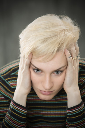 Serious head and shoulders portrait of young blonde woman in colorful striped shirt with hands on her temples.の写真素材