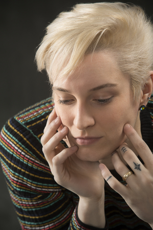 Serious head and shoulders portrait of young blonde woman in colorful striped shirt with head resting in hands.の写真素材