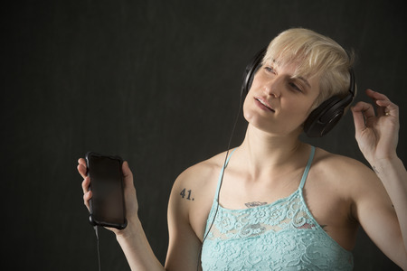 Half length portrait of young blonde woman in blue camisole with headphones on and holding a cell phone.の写真素材