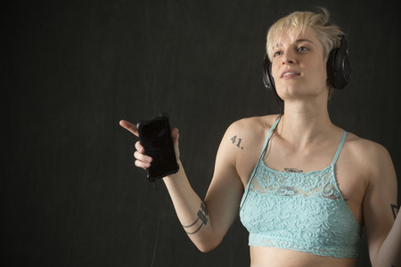 Half length portrait of young blonde woman in blue camisole with headphones on and holding a cell phone.の写真素材