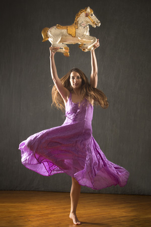 Young woman in flowing lavender dress raising overhead an antique carousel horse in the studio.の写真素材