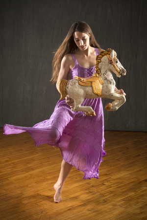 Young woman in flowing lavender dress dancing with an antique carousel horse in the studio.の写真素材