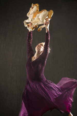 Young woman in long purple dress raising overhead an antique carousel horse in the studio.の写真素材