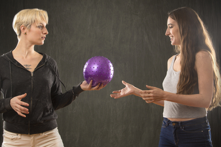 Two attractive young women friends in casual wear, playing with a purple ball in the studio against a gray background.の写真素材