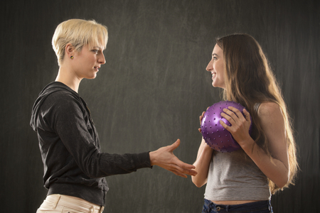 Two attractive young women friends in casual wear, playing with a purple ball in the studio against a gray background.の写真素材