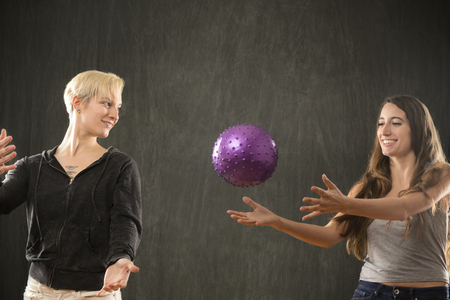 Two attractive young women friends in casual wear, playing with a purple ball in the studio against a gray background.の写真素材
