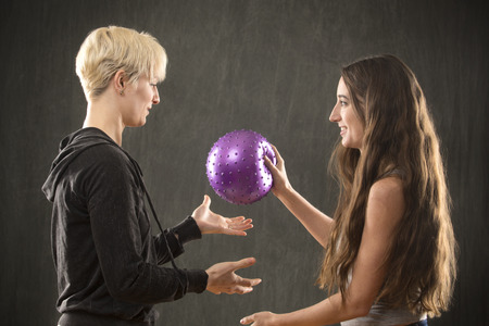 Two attractive young women friends in casual wear, playing with a purple ball in the studio against a gray background.の写真素材