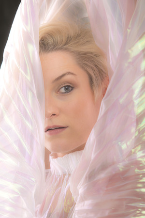 Head and shoulders portrait of a beautiful young woman wrapped in a shimmering transparent cape in the studio.の写真素材