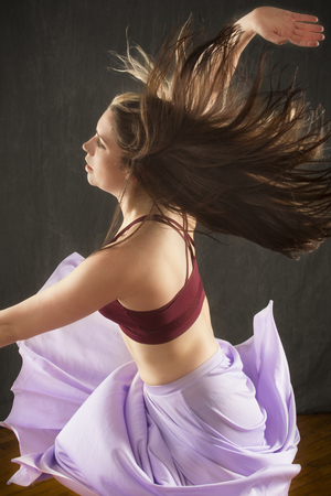 Half length studio shot of beautiful young dancer in burgundy top and lavender skirt, with arms raised while dancing in the studio.の写真素材