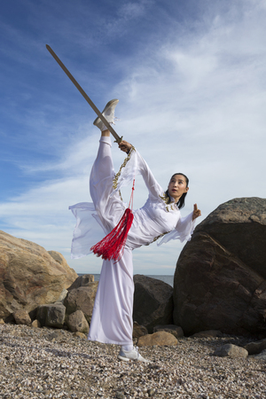 Young woman dancer in white gi performing qi gong with a sword on the rocky beach of Hammonasset State Park in Madison, Connecticut.の写真素材