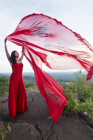 Female dancer in red dress, waving into the wind in late afternoon on the mountain, a red fabric with Chinese calligraphy on it, at Penwood State Park in Bloomfield, Connecticut.の写真素材