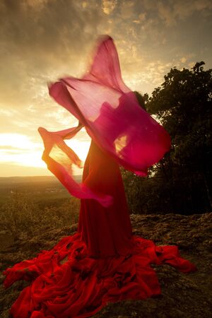 Female dancer in red dress, waving into the wind at sunset on the mountain a red fan with flowing fabric on it, at Penwood State Park in Bloomfield, Connecticut.の写真素材