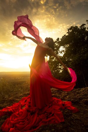Female dancer in red dress, waving into the wind at sunset on the mountain a red fan with flowing fabric on it, at Penwood State Park in Bloomfield, Connecticut.の写真素材