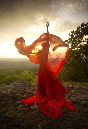 Female dancer in red dress, waving into the wind at sunset on the mountain a red fabric with Chinese calligraphy on it, at Penwood State Park in Bloomfield, Connecticut.の写真素材
