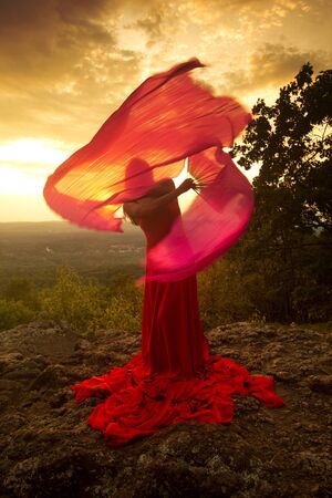 Female dancer in red dress, waving into the wind at sunset on the mountain a red fan with flowing fabric on it, at Penwood State Park in Bloomfield, Connecticut.の写真素材