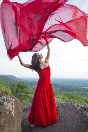 Female dancer in red dress, waving into the wind in late afternoon on the mountain, a red fabric with Chinese calligraphy on it, at Penwood State Park in Bloomfield, Connecticut.の写真素材