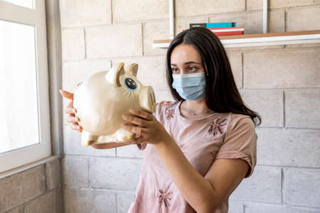 Young woman with face mask, worried face looking at his piggy bank in times of pandemic by COVID-19.の写真素材