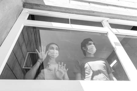 Man and woman in quarantine with face masks looking out of their house through the window. Black and...