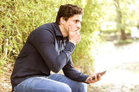 Young troubled businessman sitting in garden with cell phone in his hand.
