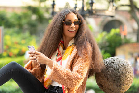 Beautiful latin woman with sunglasses, smiling happy with her cellphone in outdoor park