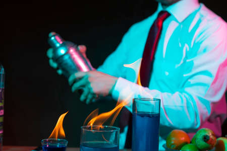 Bartender preparing alcoholic drink with a cocktail shaker at the bar with some glasses of tequila on fire. Dramatic lightsの写真素材