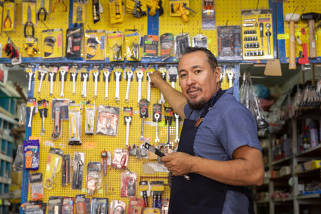 A handsome adult businessman wearing an apron, with a joyful expression, showing at camera products from his business full of hardware products.の写真素材