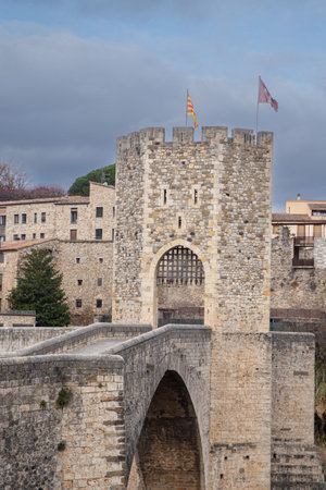 Old entrance to Rupit, a beautiful medieval town in Spain preserved to this day as a tourist destination with its imposing castle-shaped entrance built of stone and its solid steel gate and flags at the top and in the distance you can see the houses and what appears to be a chapelの写真素材