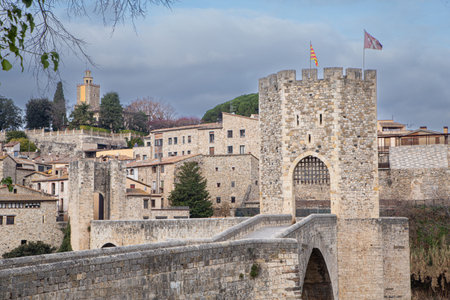 Old entrance to Rupit, a beautiful medieval town in Spain preserved to this day as a tourist destination with its imposing castle-shaped entrance built of stone and its solid steel gate and flags at the top and in the distance you can see the houses and what appears to be a chapelの写真素材