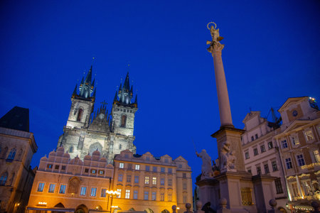 Medieval castle decorated with an antique clock, saints and beautiful towers located in the center of the modern and bustling city of Prague, between streets busy with modern transportation surrounded by beautiful buildings, houses and a monument illuminated at nightの写真素材