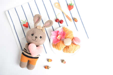 Cute crochet brown bunny eating healthy breakfast meal with croissants, Macaron, strawberry, dried acorn, spoon, pink orchid, cherry, flat lay, top view, food and beverage in white backgroundの写真素材
