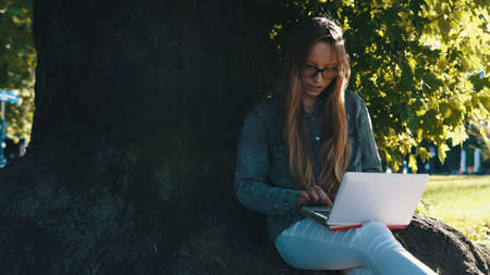 Young caucasian woman working on the laptop sitting near the tree in the park. High quality photoの写真素材